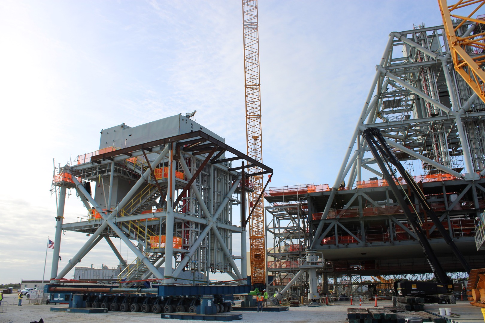 Large yellow cranes and other equipment position a gray rocket launch module prior to erection atop previously installed modules.